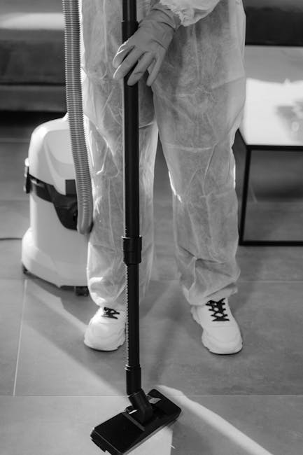 A woman with curly hair, dressed in a beige jacket, dark jeans, and white sneakers, is operating a yellow vacuum cleaner on a multicolored patterned carpet in a living room. She is holding the vacuum'
