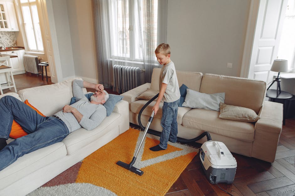 Close-up of a person vacuuming a patterned area rug with a black and gray vacuum cleaner nozzle, in a well-lit room, demonstrating surface cleaning and deep cleaning techniques. The room features hard